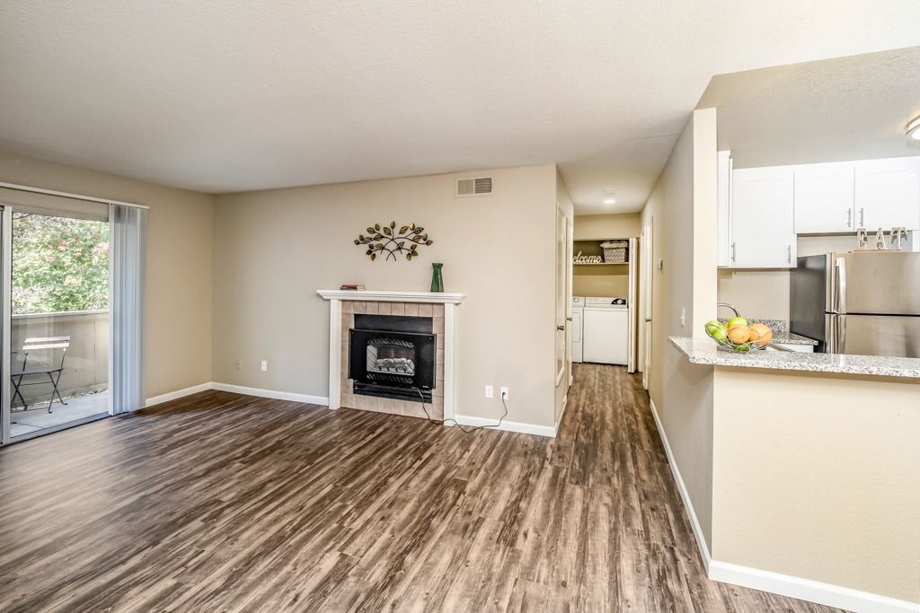 Living area with a fireplace, wood-like flooring, and open kitchen in an apartment.