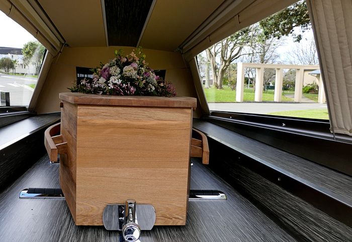 Bara di legno in un carro funebre, sormontata da fiori, vista dal portellone posteriore aperto.
