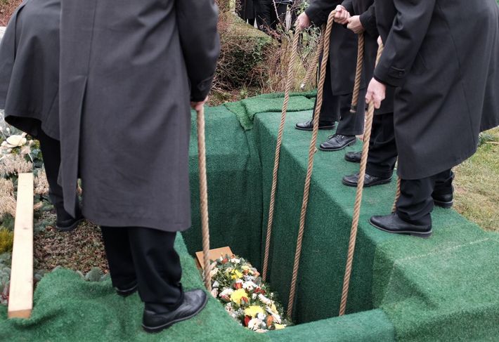 Persone con indosso cappotti scuri sono in piedi intorno a una tomba con un'apertura piena di fiori e ricoperta di erba verde.