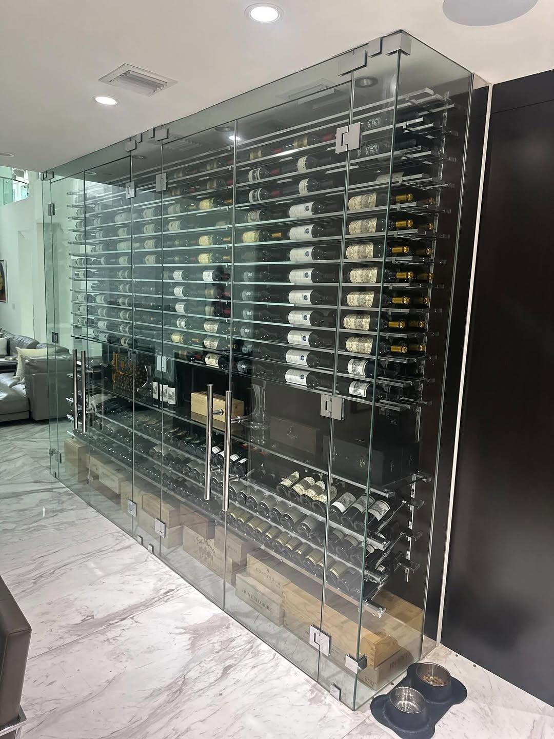 Glass wine display cabinet in a modern room with shelves of bottles and a marble floor.