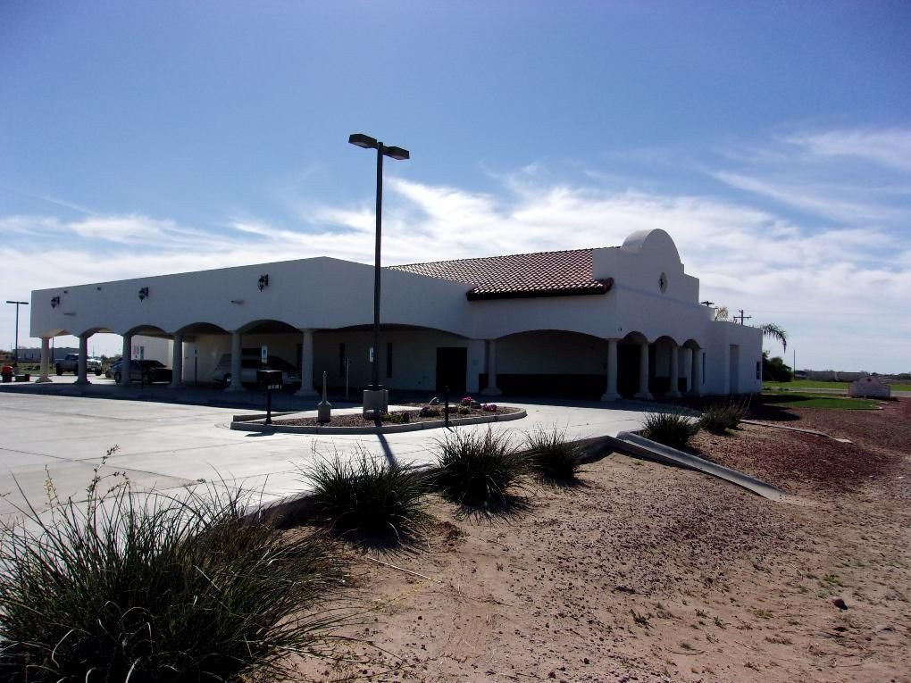 White commercial building with arched windows and a sign, beside a parking lot and desert landscaping under a blue sky