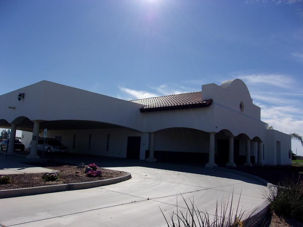 White stucco building with arched entrance and red tile roof under a bright sunny sky.