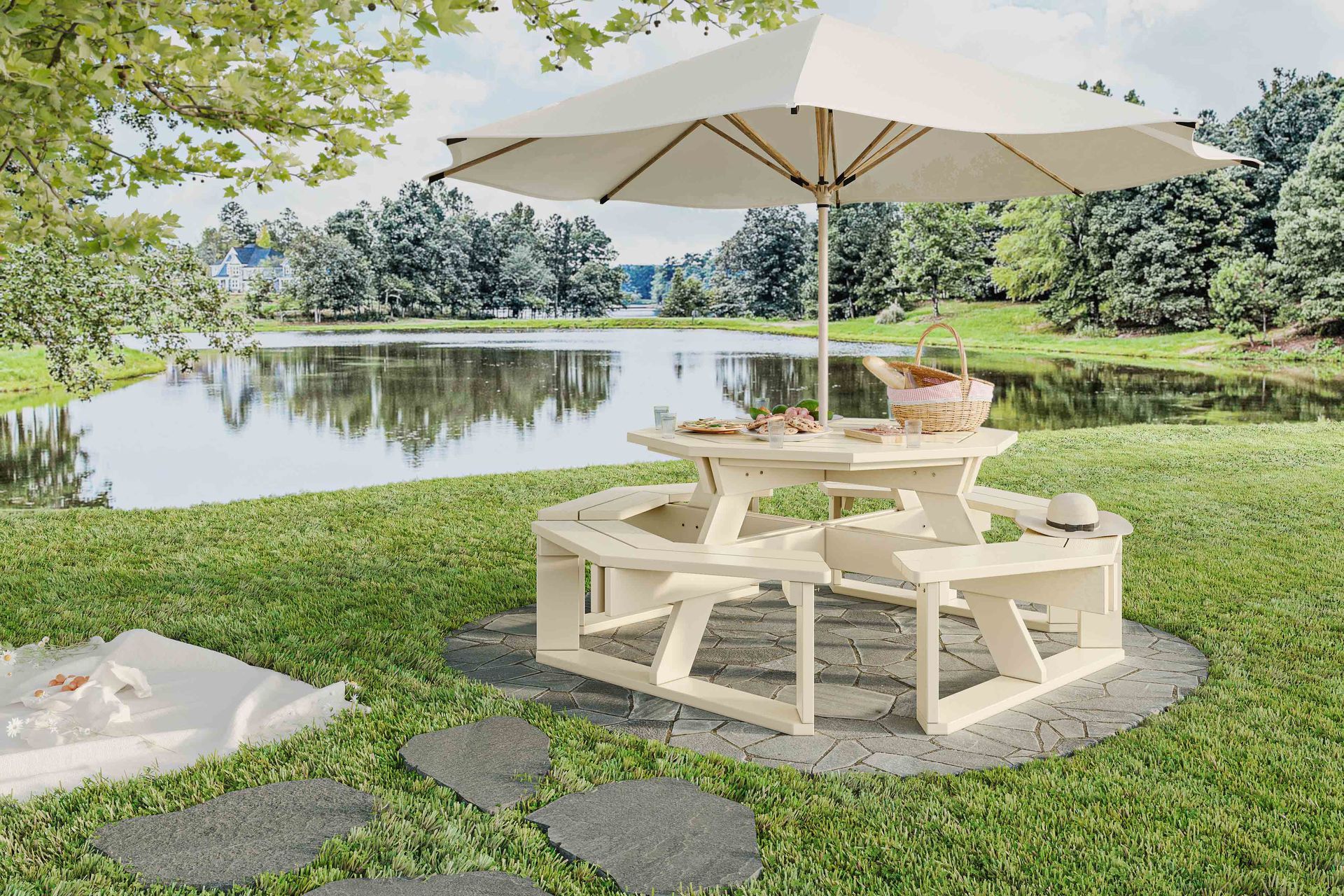 Picnic table set by a pond, shaded by an umbrella. Basket and food on table. Green grass.