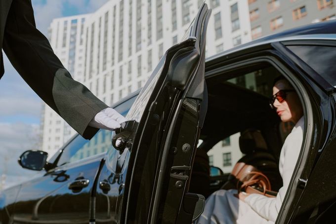 Driver in sunglasses exits a black car while a gloved hand holds the door open.