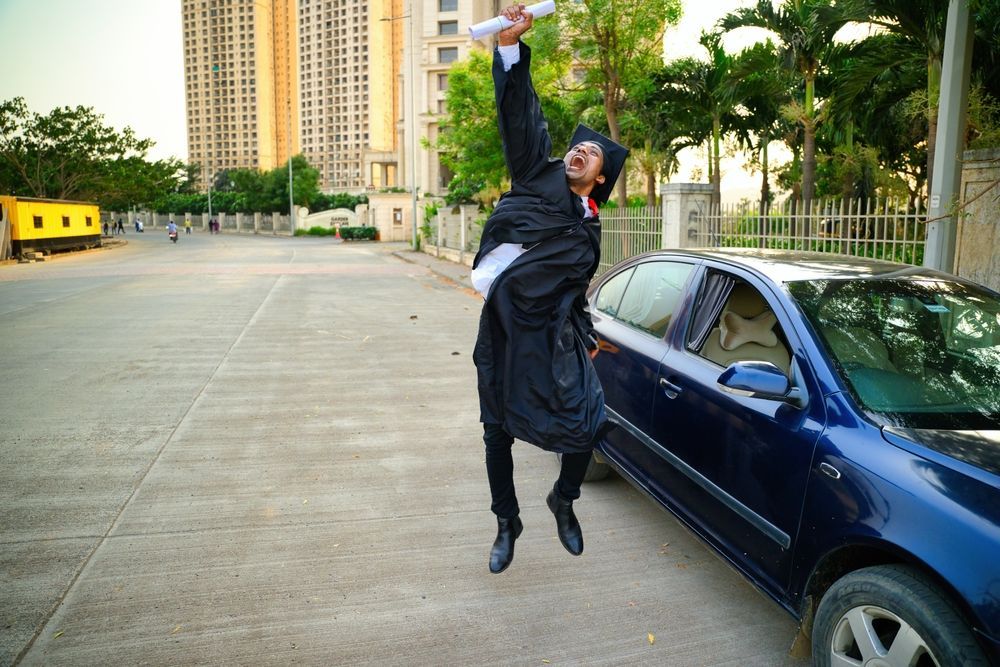 Man in dark clothes mid-jump beside a blue car on a city street with tall buildings behind him