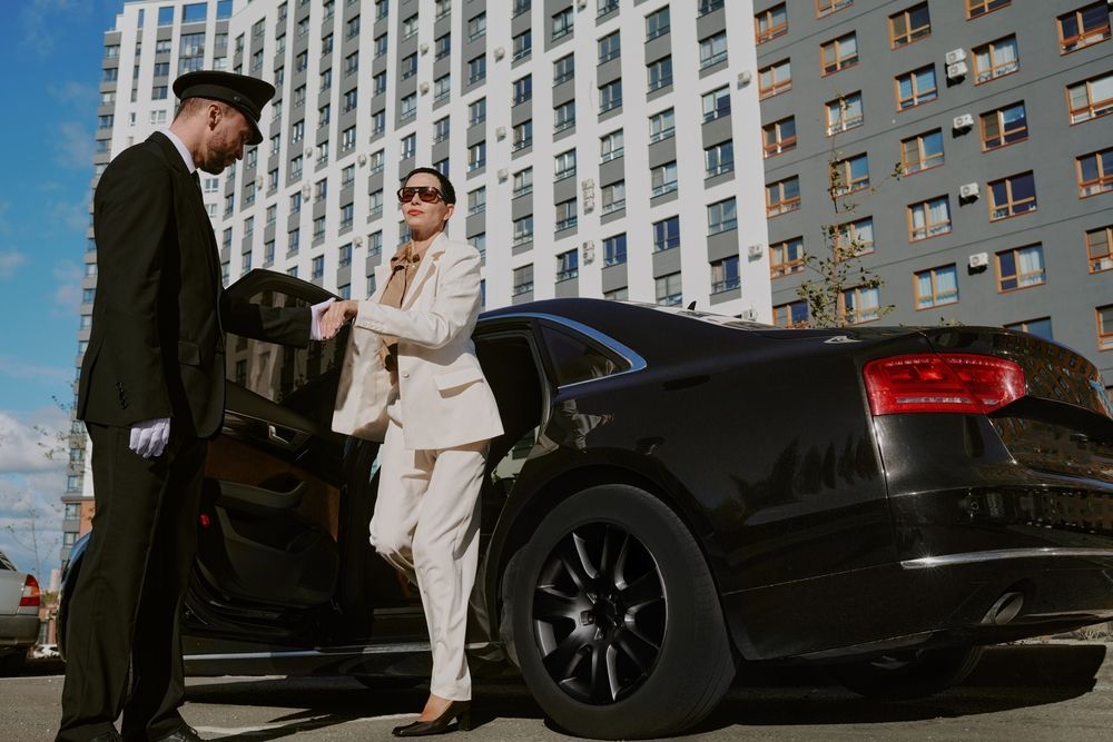 Two people stand by an open black sedan in front of tall city buildings.