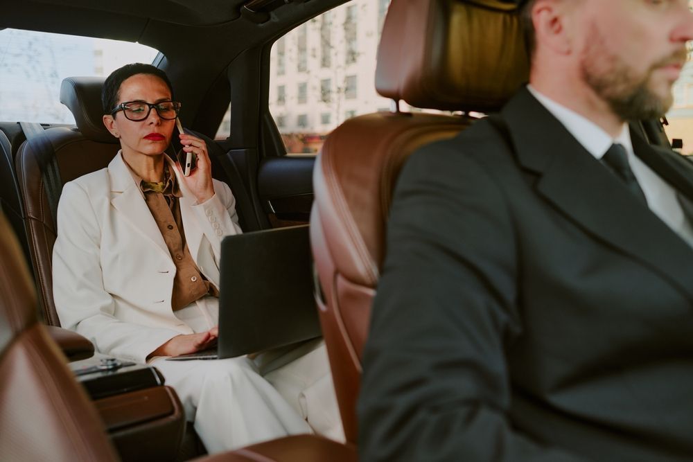 Two professionals in a car, one on a laptop in backseat while the driver focuses ahead.