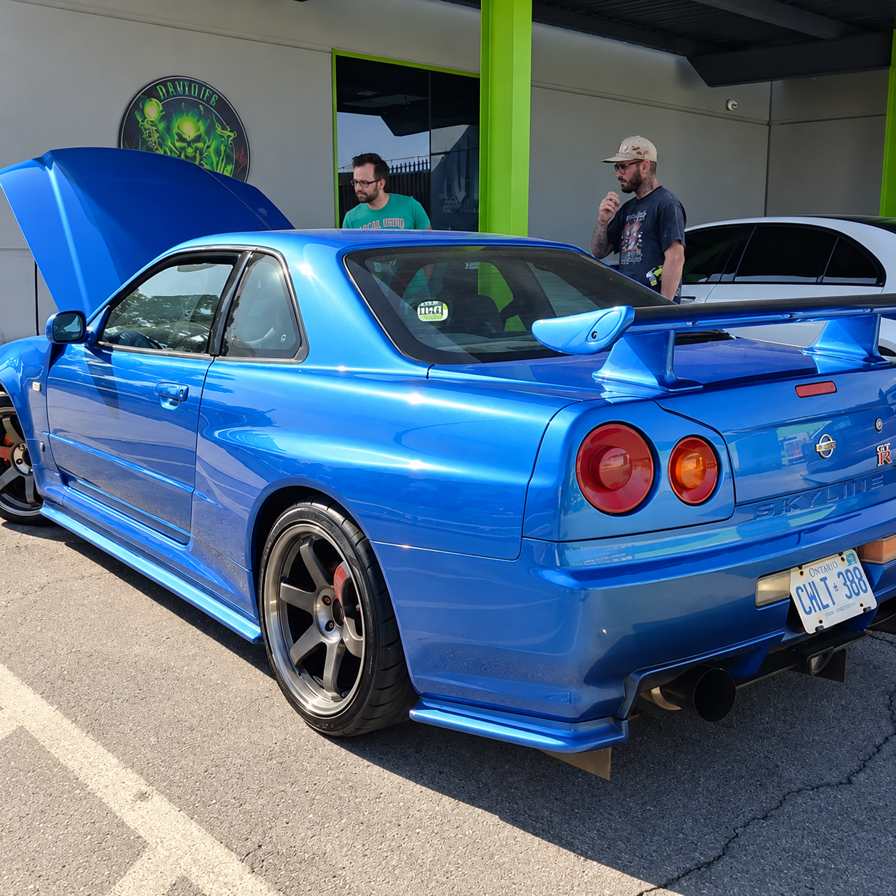 Blue sports coupe with rear spoiler at a car meet, hood open, two people standing nearby