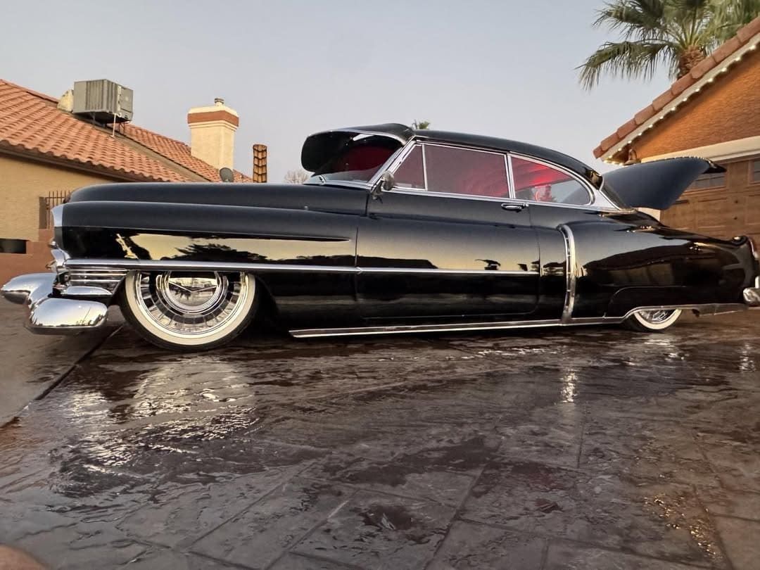 Classic black lowrider car parked on a wet driveway in front of a house
