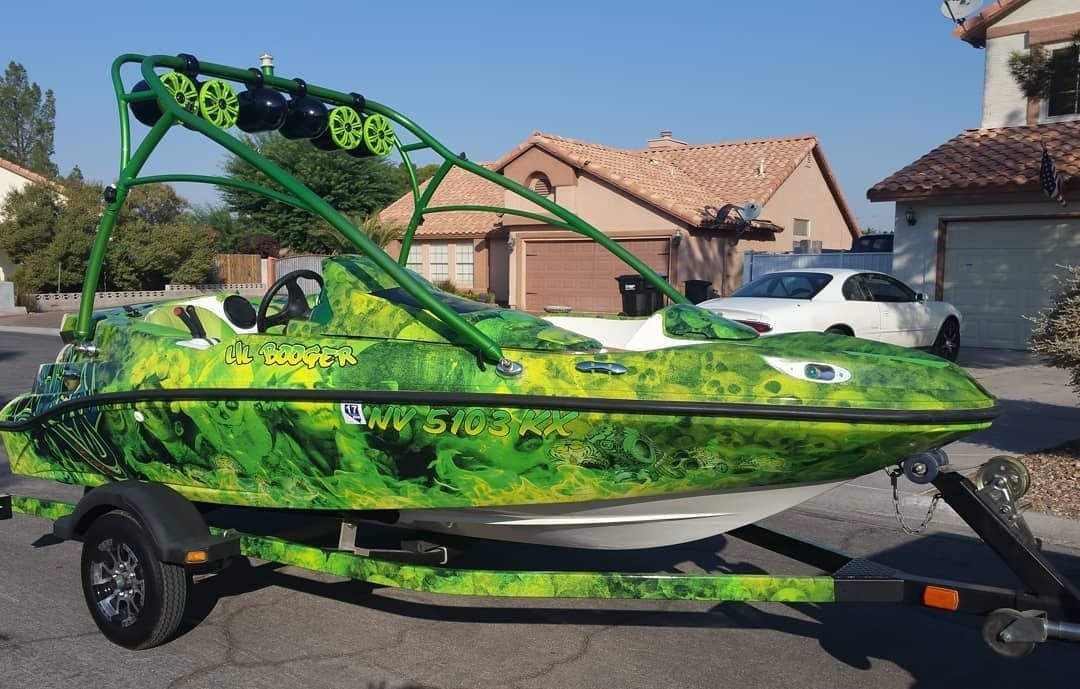 Green speedboat on a trailer parked in a suburban driveway with houses in the background
