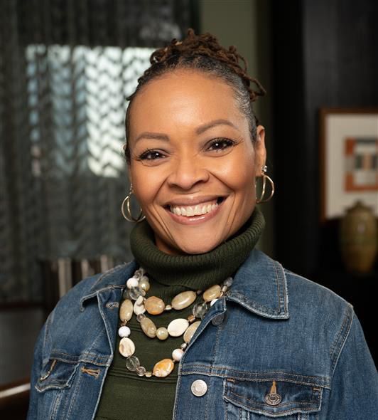 Woman smiles brightly, wearing denim jacket over green turtleneck and beaded necklace.