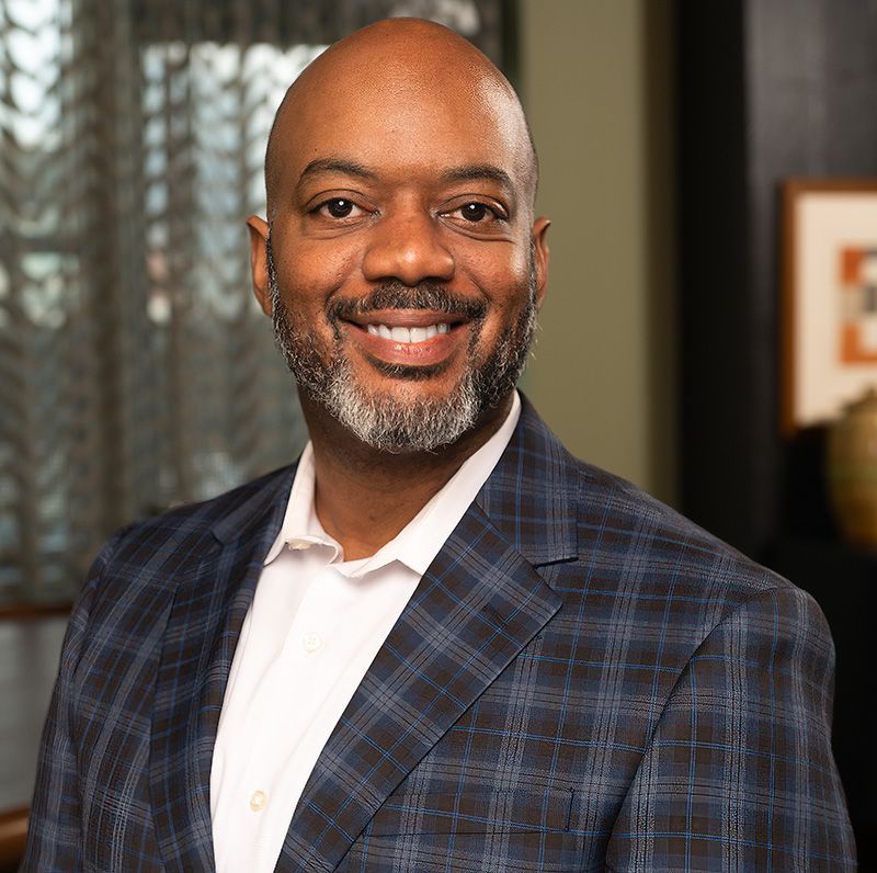 Smiling Black man in blue plaid blazer, white shirt. Indoor setting, office.