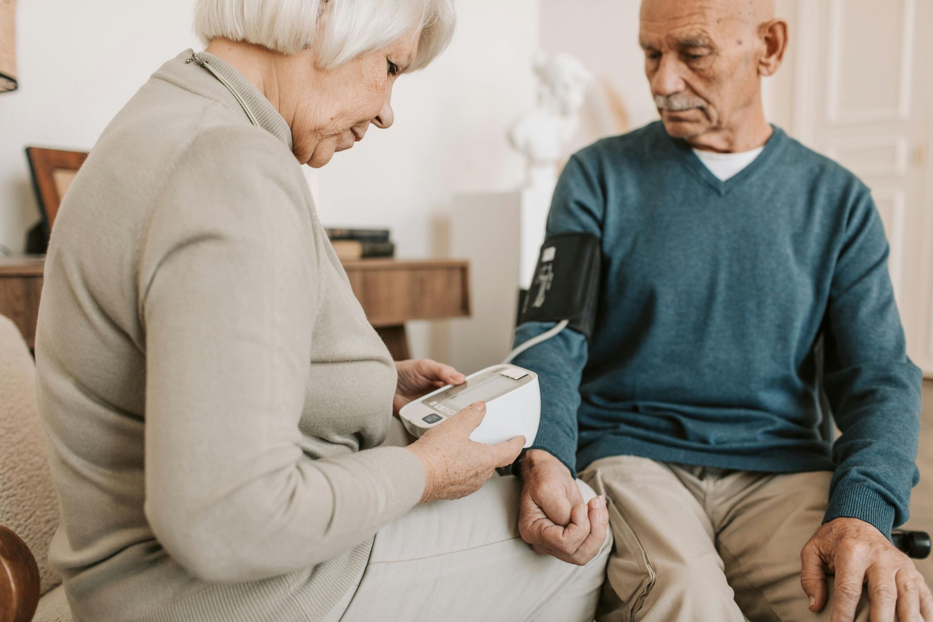 Woman taking man's blood pressure in a living room. Both are elderly.