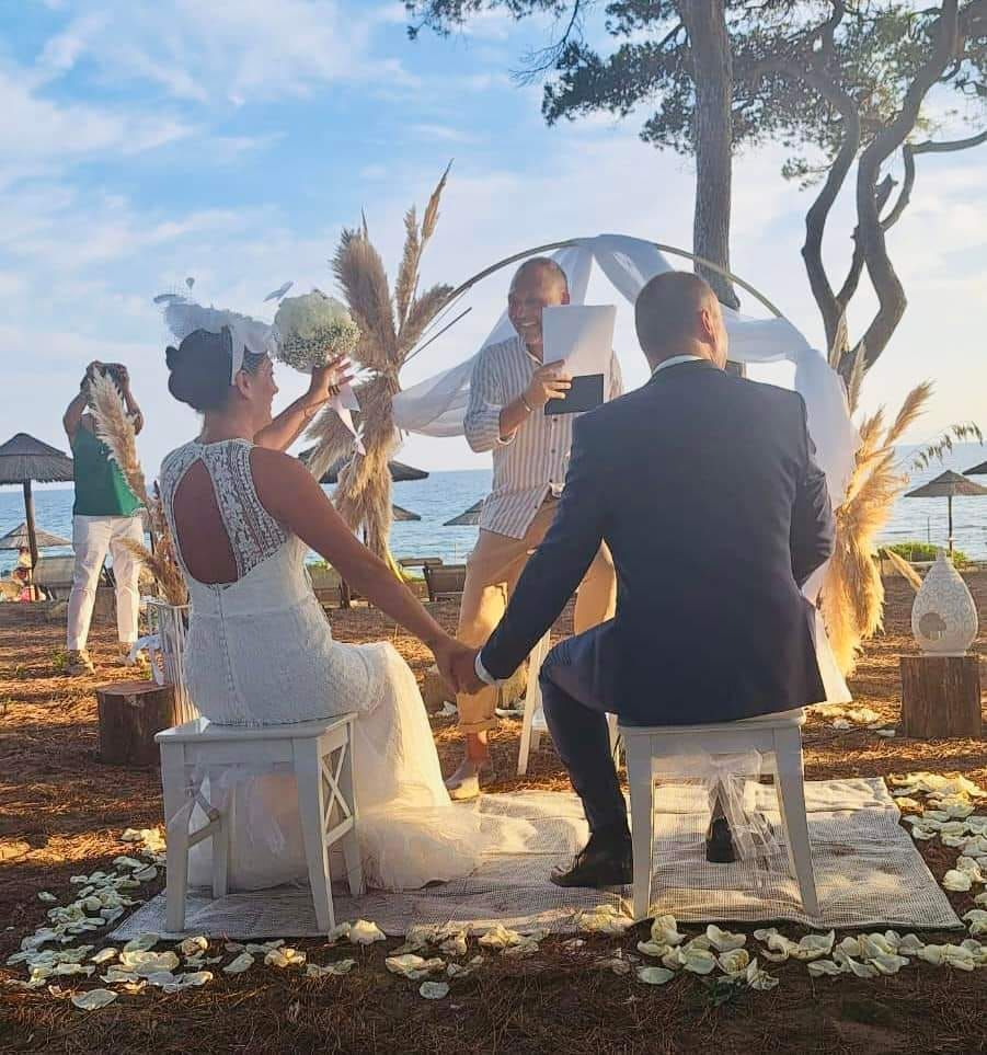 Cerimonia nuziale sulla spiaggia; coppia che si tiene per mano, celebrante che legge, arco decorativo.