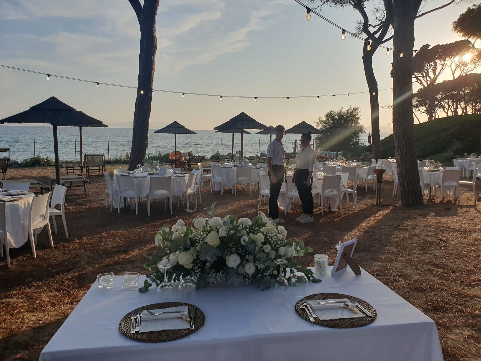 Tavoli per il ricevimento di nozze allestiti vicino alla spiaggia al tramonto, con centrotavola floreale.