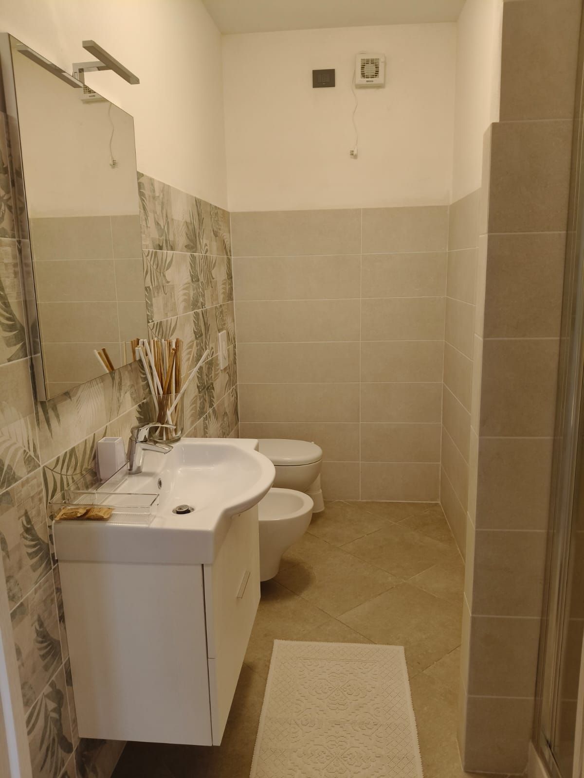 Bagno con lavandino e WC bianchi, pareti e pavimento piastrellati beige e parete decorata dietro il lavandino.