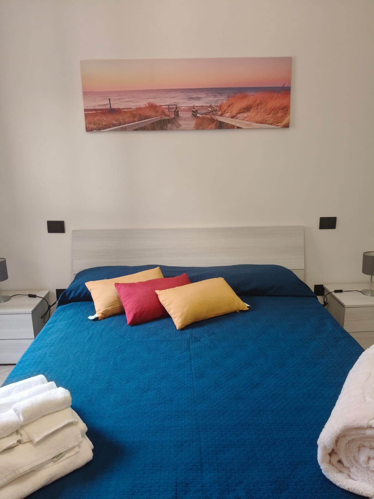 Camera da letto con copriletto blu, cuscini, asciugamani e un dipinto raffigurante un paesaggio marino sopra il letto.