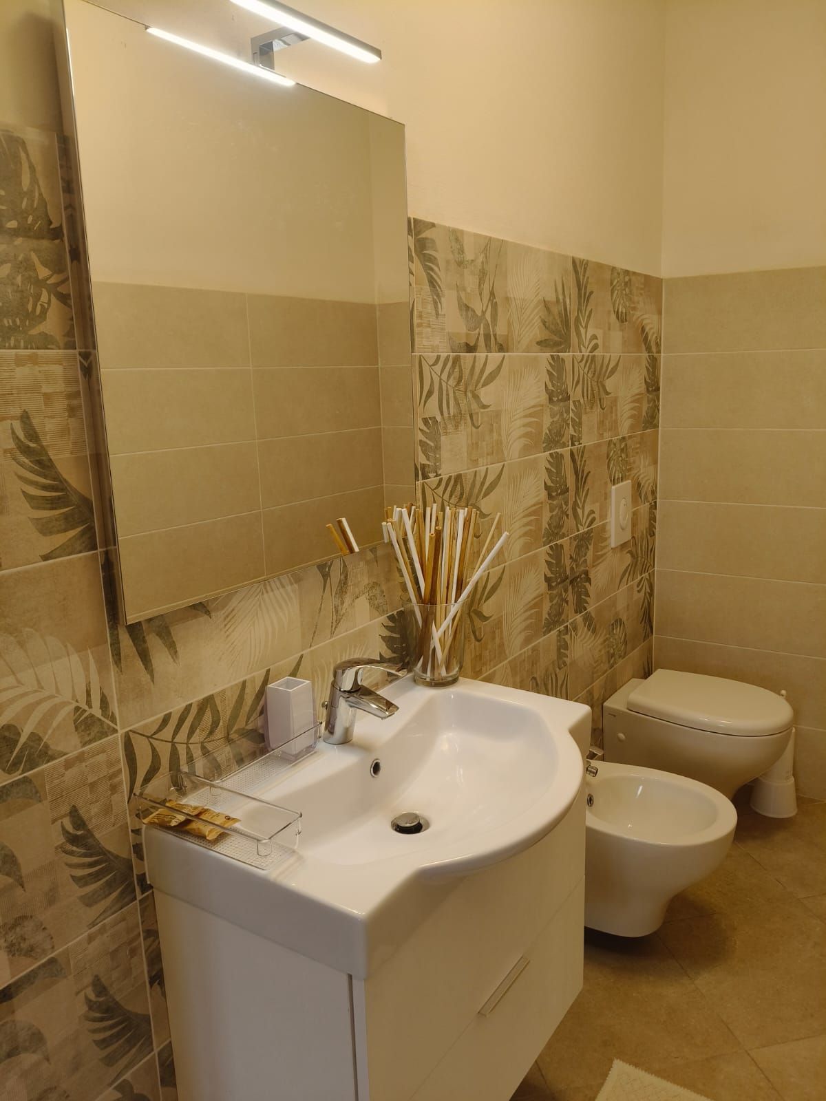 Bagno con lavabo, WC e bidet bianchi. Pareti con piastrelle beige e fantasia, specchio, diffusore a bastoncini.