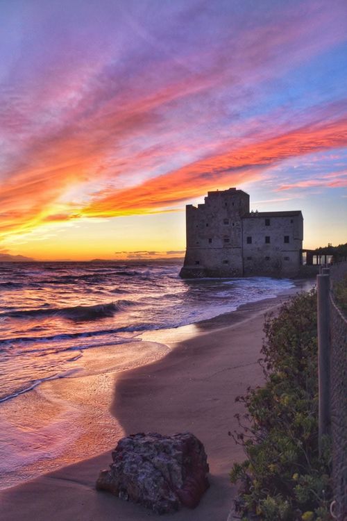 Vista costiera: torre in pietra su una spiaggia, onde che si infrangono sulla riva sotto un cielo azzurro.