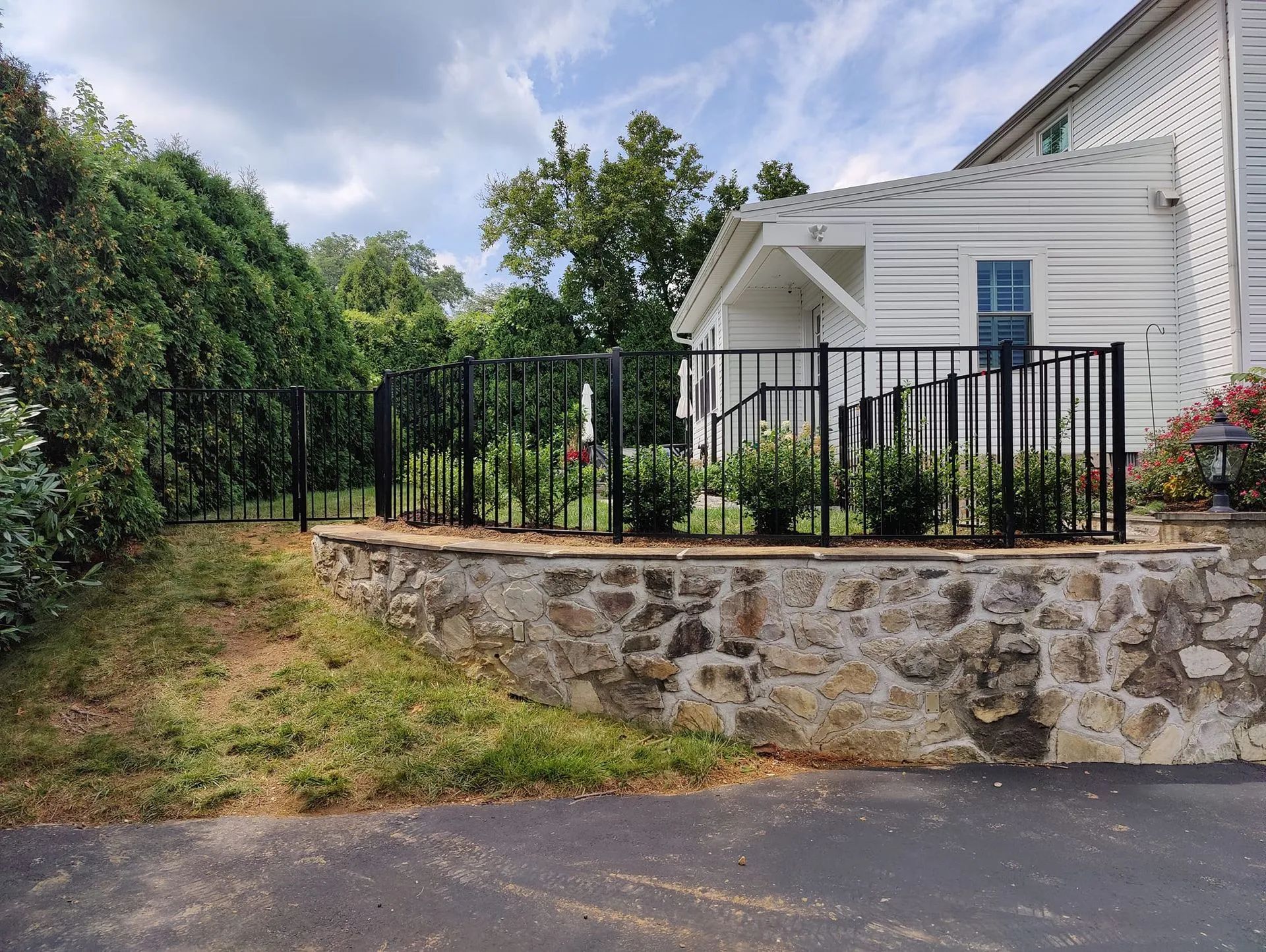 White house with black metal fence and stone retaining wall beside a grassy roadside.