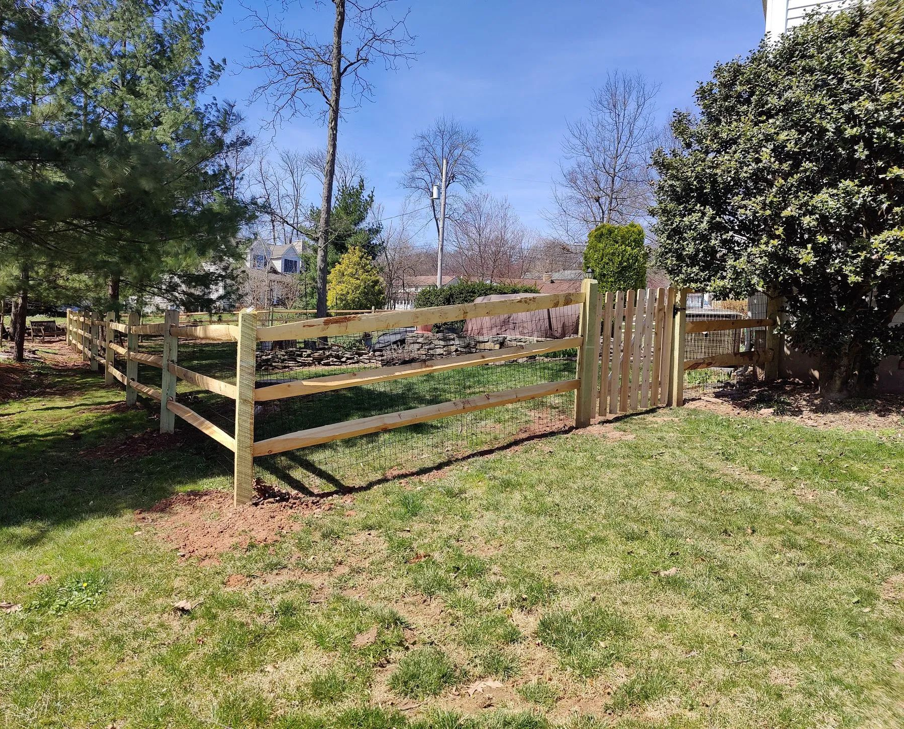Wooden fence gate in a sunny grassy yard with trees and a stone wall beyond