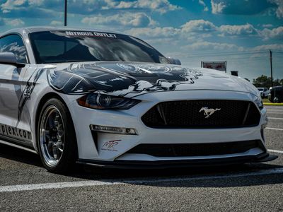 A white Ford Mustang with a black graphic on the hood parked in an outdoor lot under a cloudy blue sky.