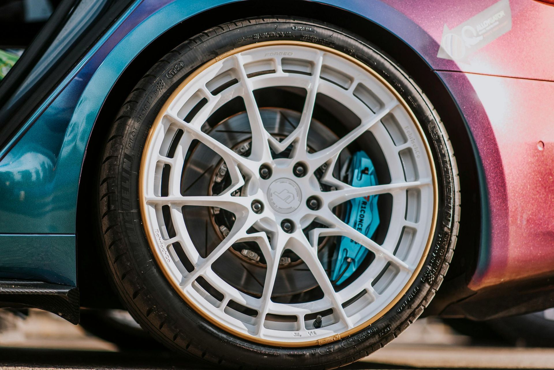 A close-up of a white, multi-spoke car wheel with a gold rim and a bright blue brake caliper on a two-tone car.