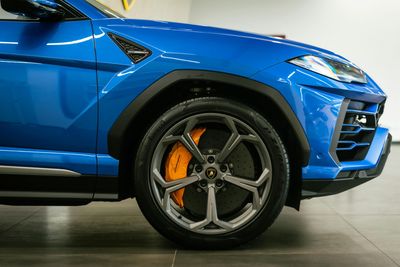 Close-up profile of a bright blue Lamborghini Urus SUV, highlighting the front wheel, orange brake caliper, and design.