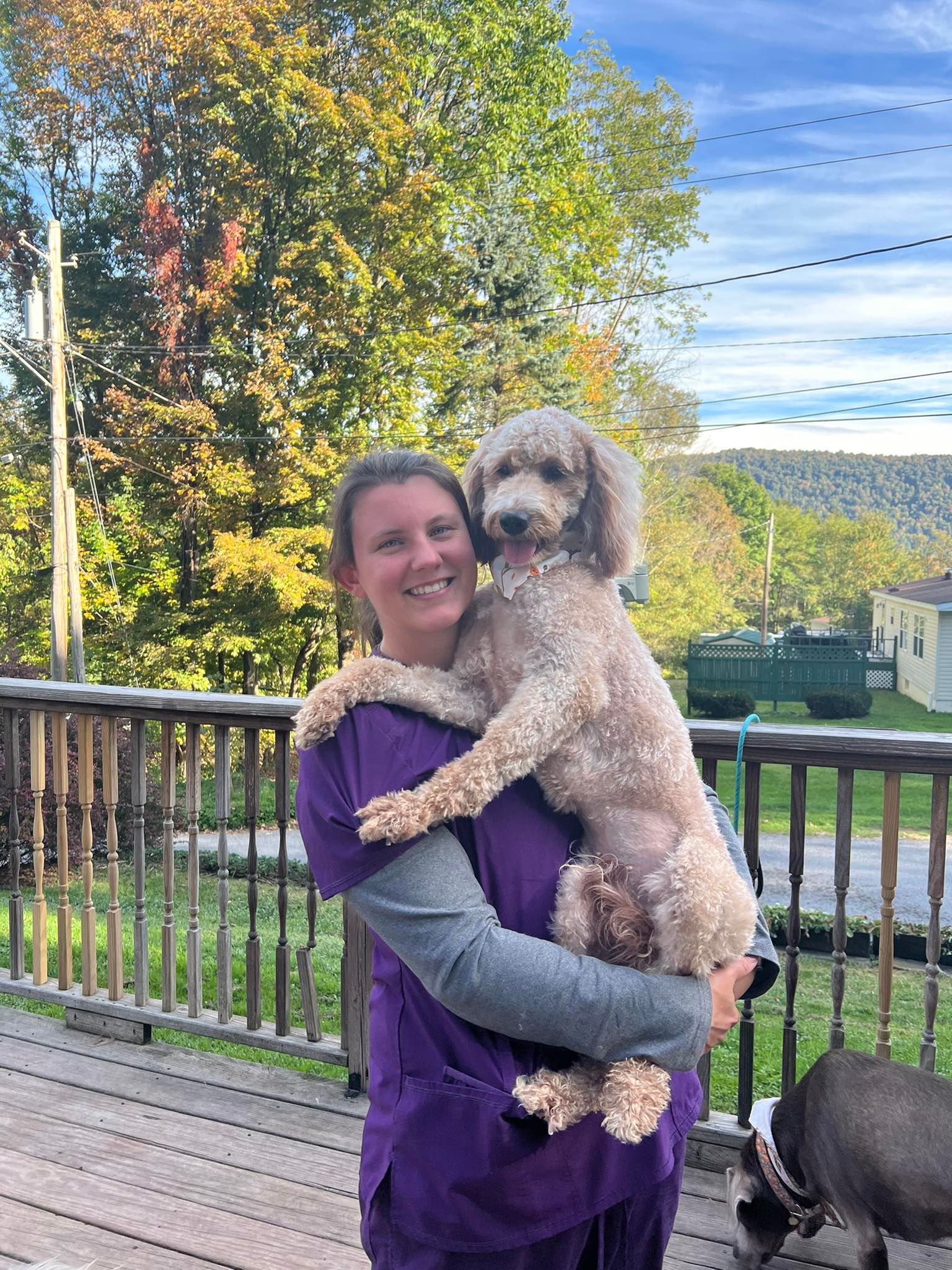 Woman in purple scrubs holding a poodle on a deck, smiling. Green and yellow trees, blue sky in the background.