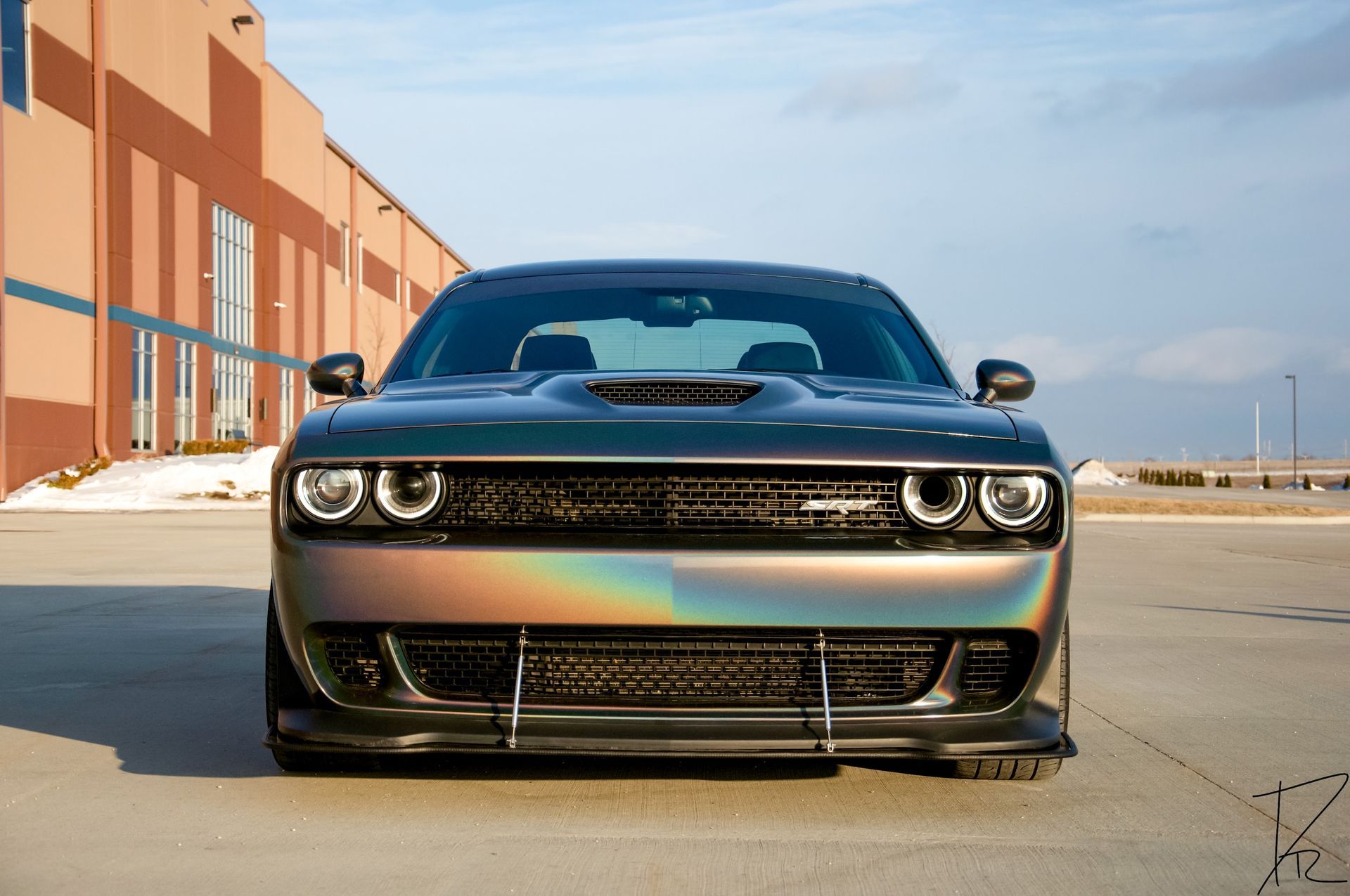 Front view of a black Dodge Challenger parked on a sunlit street beside brick buildings.