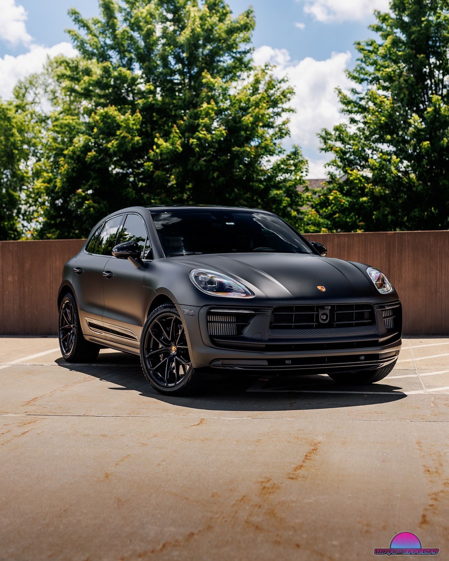 Black Porsche SUV parked outdoors in sunlight, with trees and a wooden fence in the background.