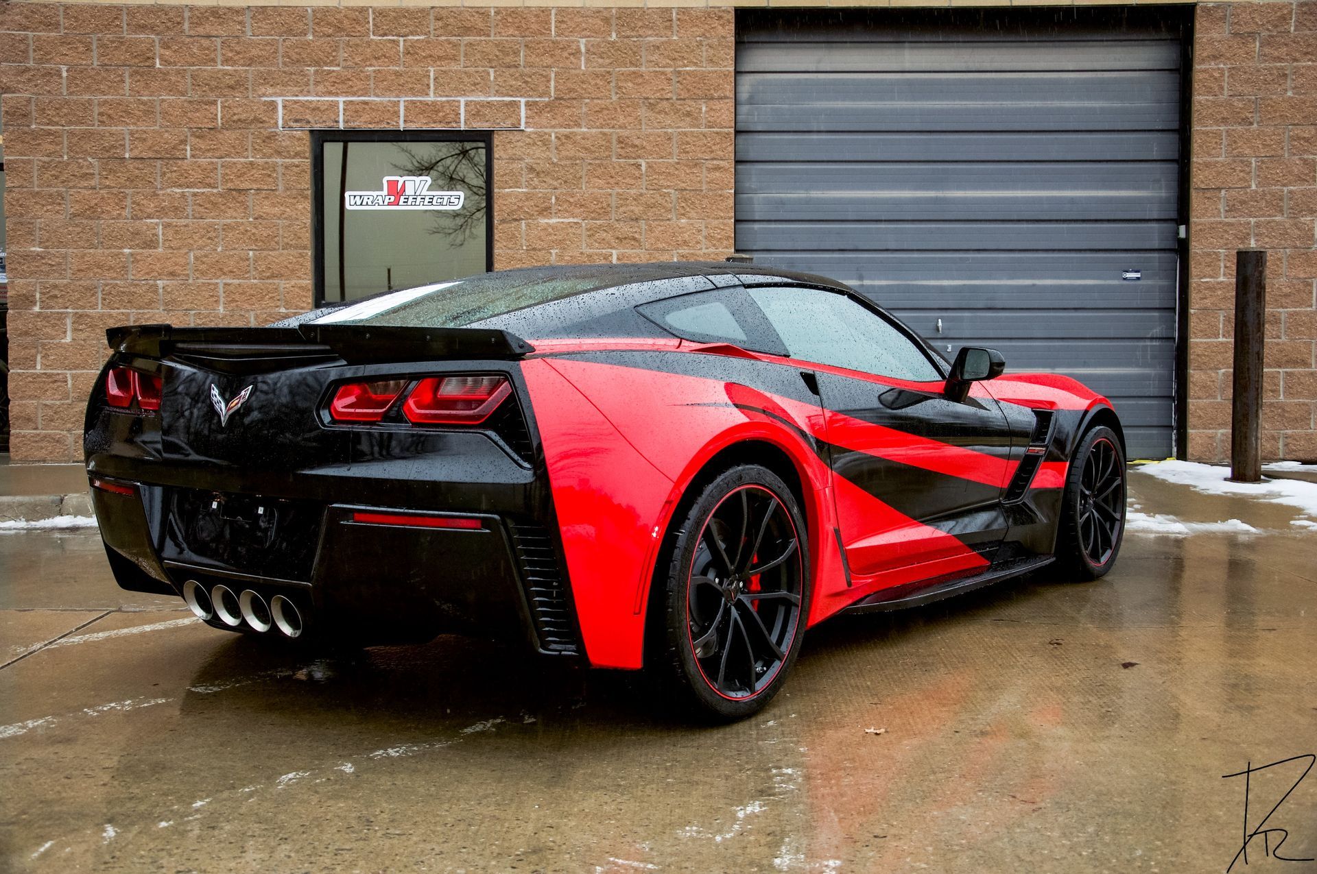 Red and black sports car parked on wet pavement beside a brick building and garage door.