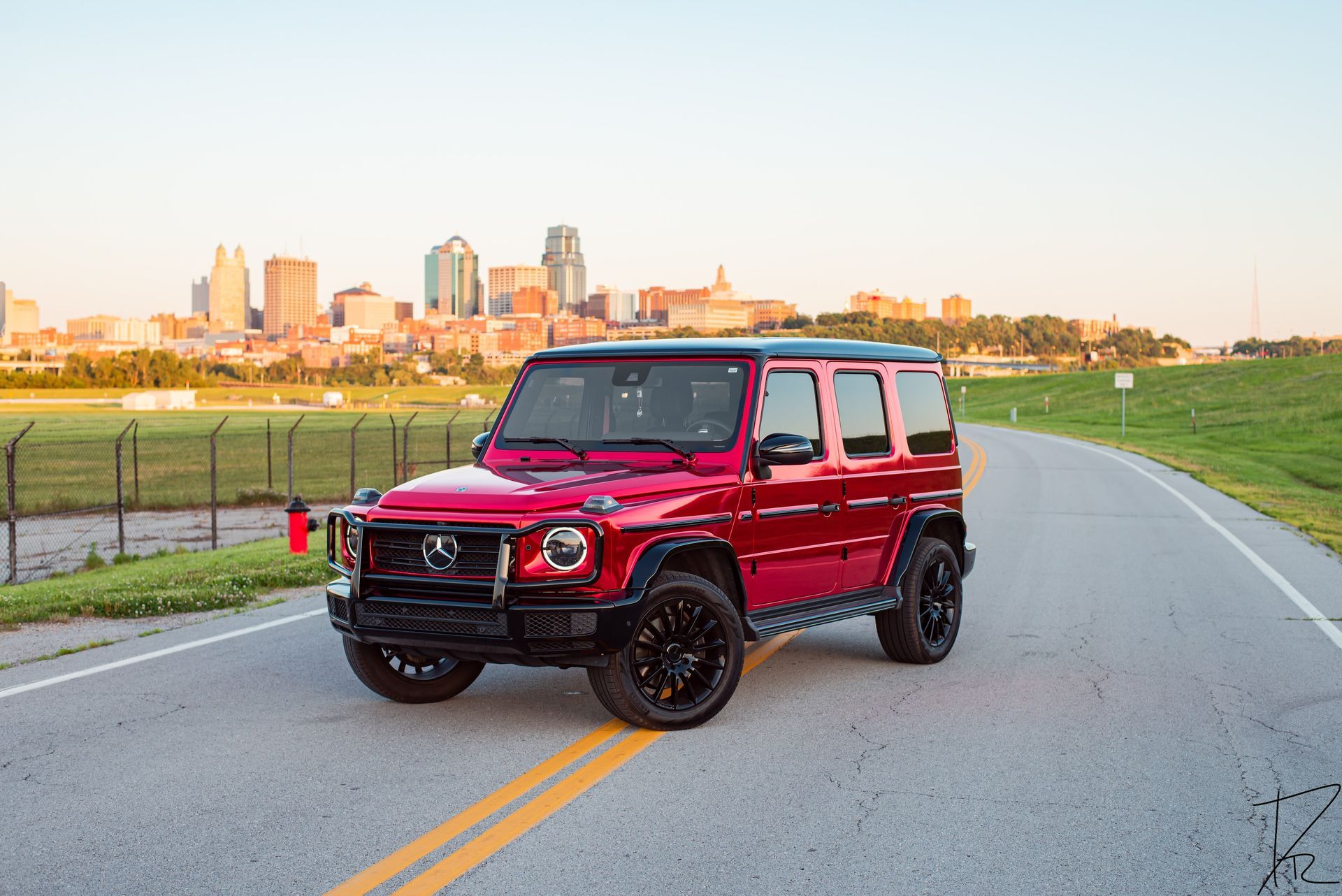 Red SUV parked on a road beside a grassy field with a city skyline in the background.