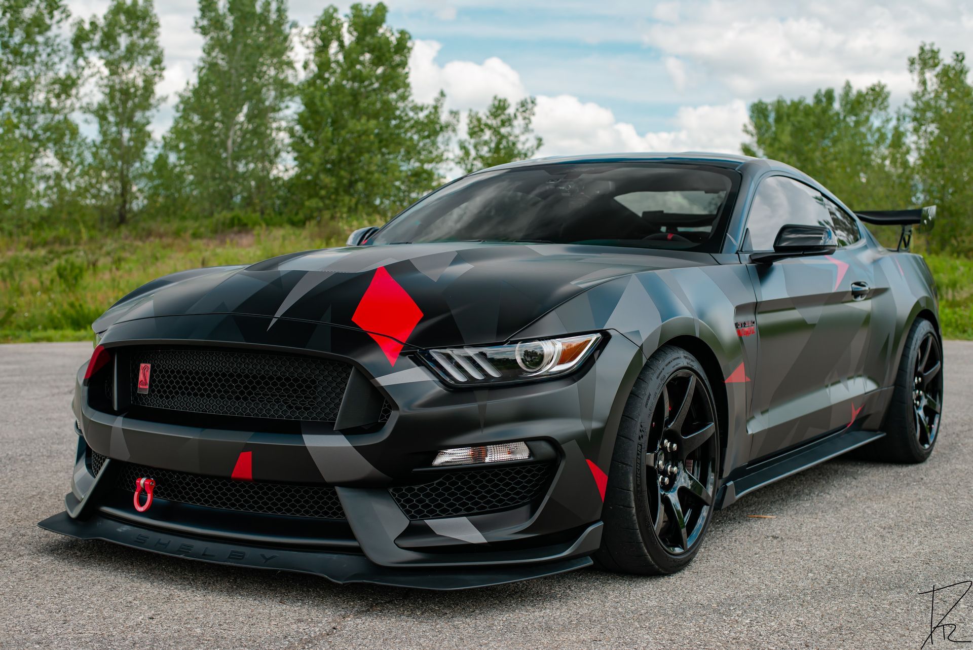Black sports car with red accents parked on a road beside trees and water under a cloudy sky