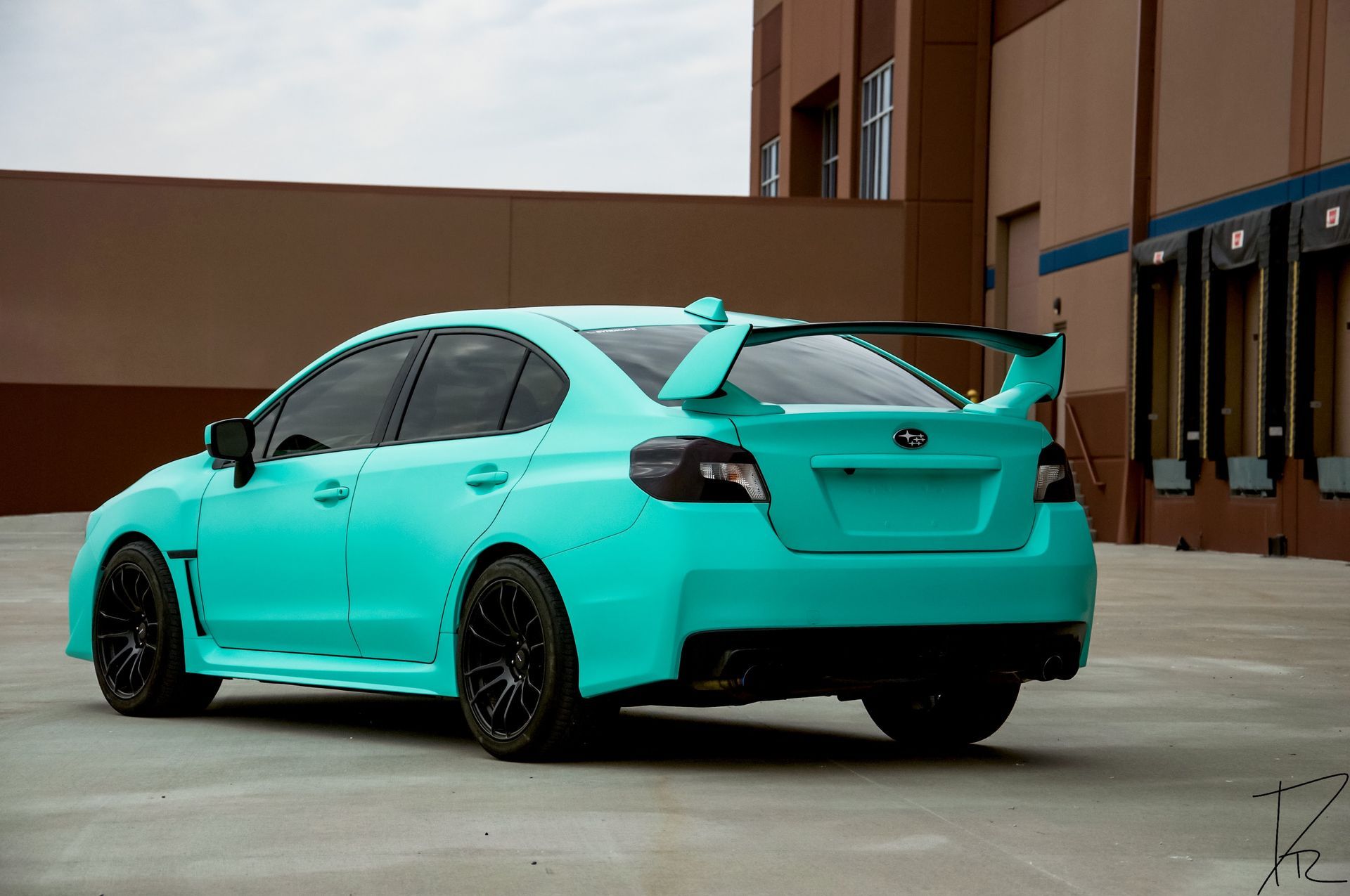 Turquoise sports sedan parked outdoors beside a building, rear three-quarter view.