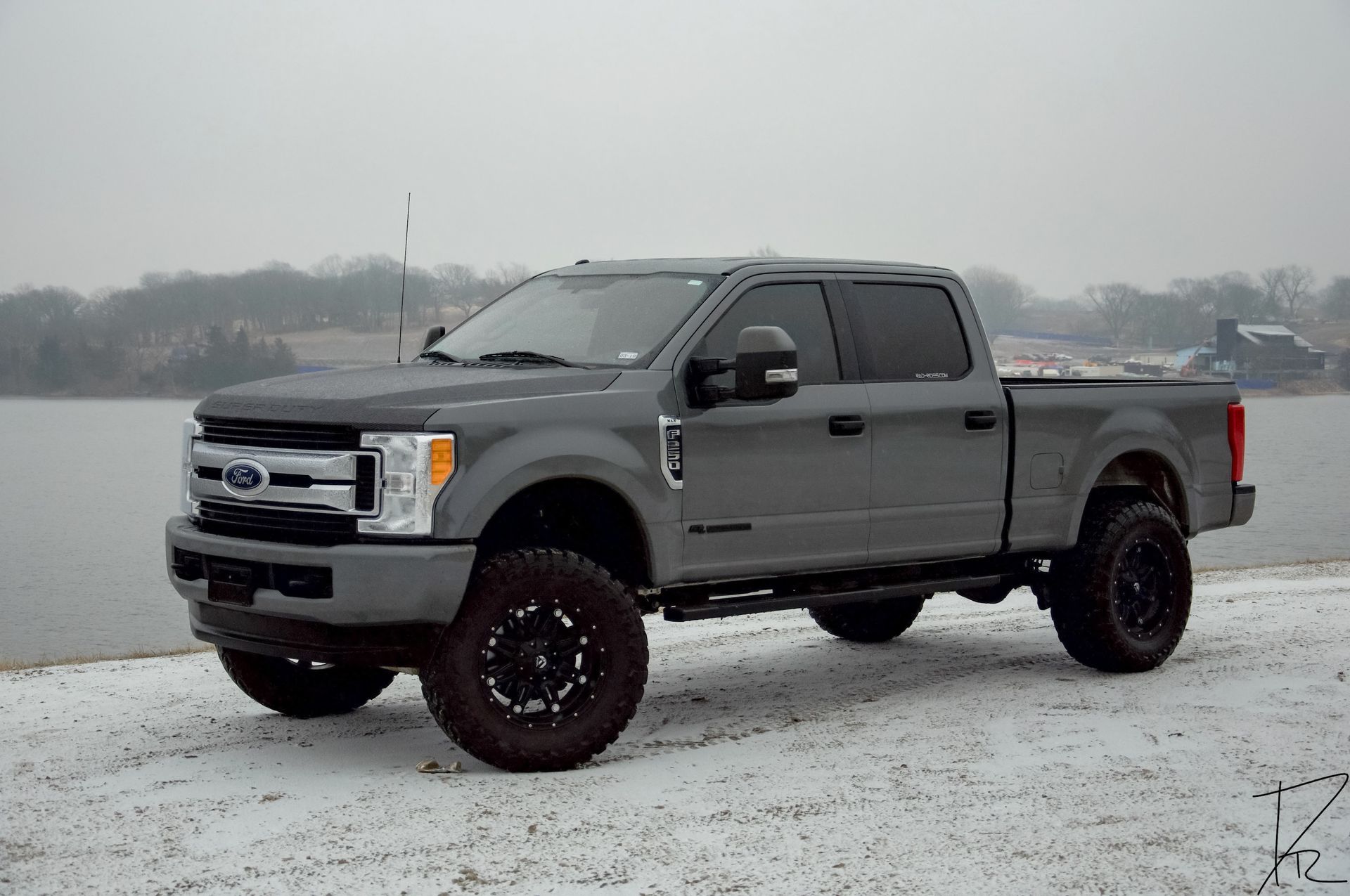 Gray lifted Ford Super Duty pickup parked on a snowy lakeshore under an overcast sky