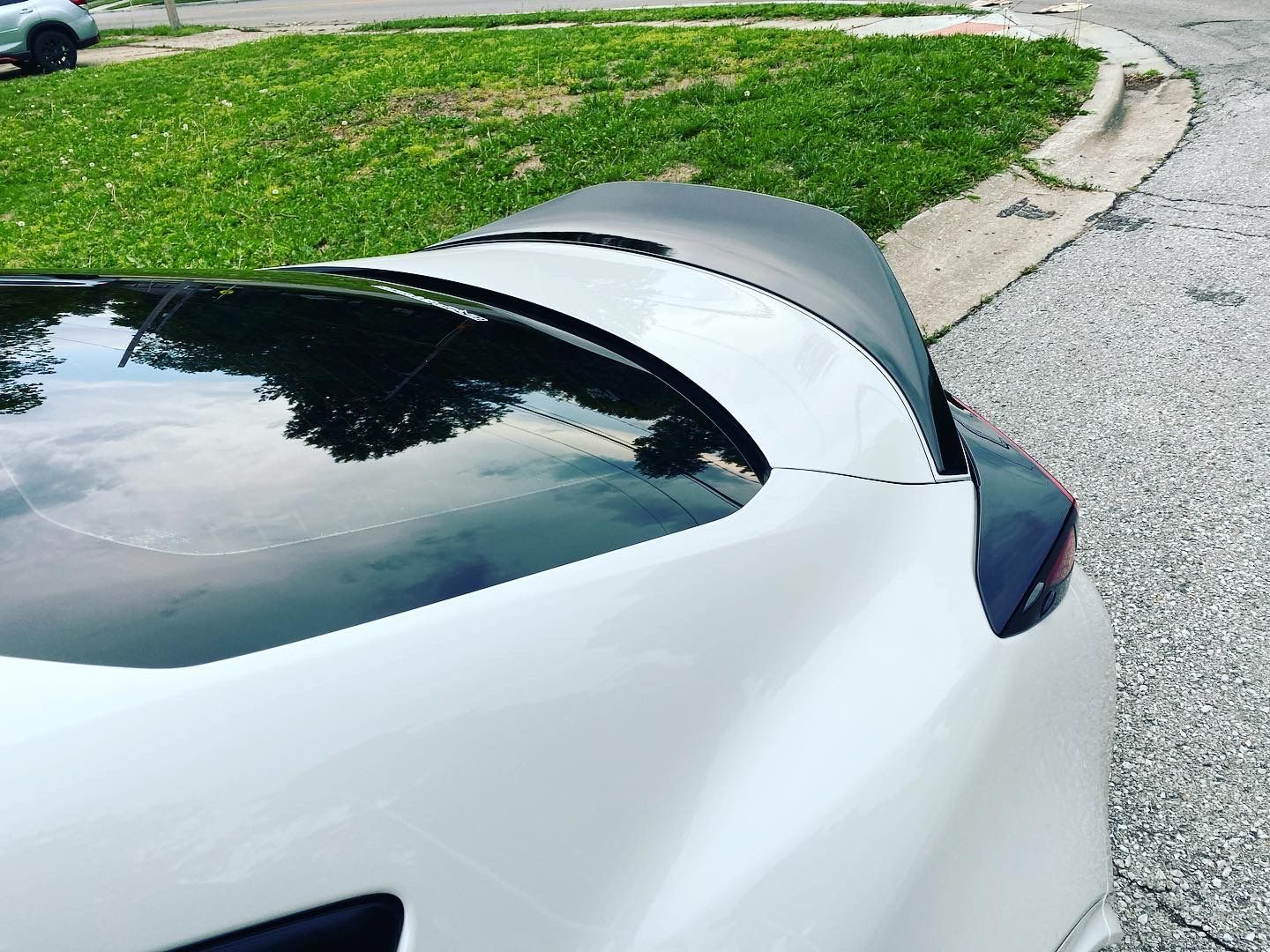 White sports car with a raised black rear spoiler parked beside a grassy curb