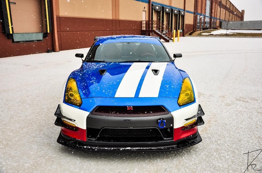 Blue sports car with white racing stripes parked in a snowy lot beside brick buildings