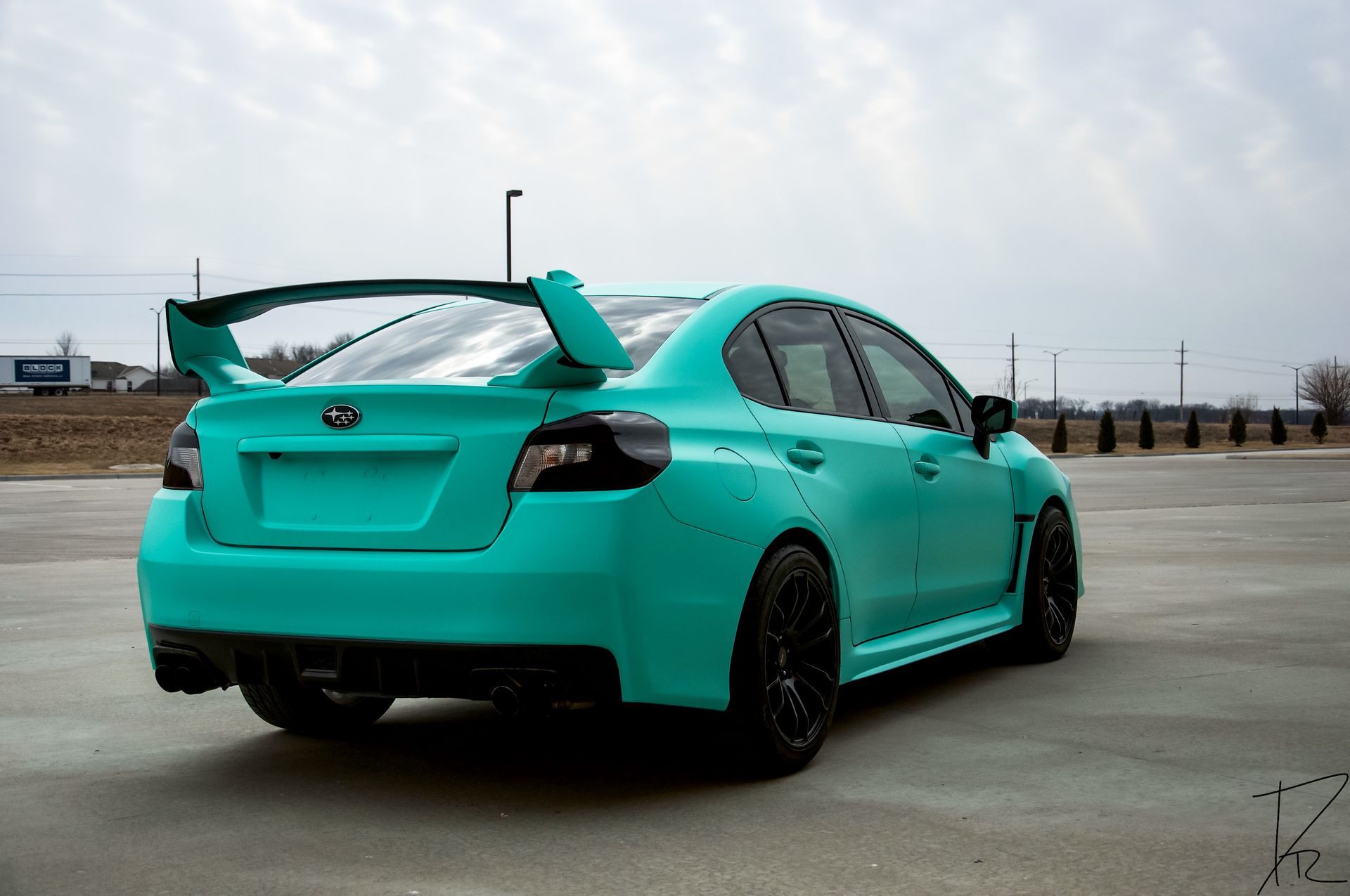 Turquoise modified sports sedan with rear spoiler parked on a concrete lot under a cloudy sky
