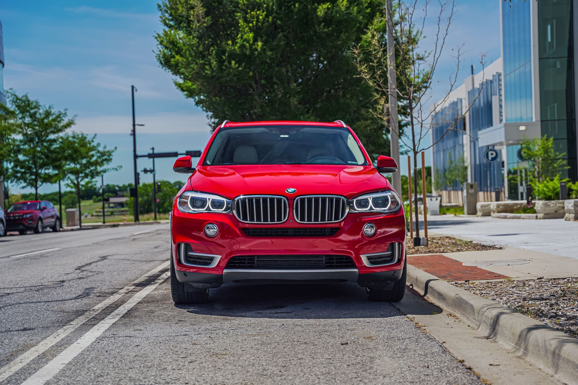 Red SUV parked on a street beside a sidewalk and modern buildings on a sunny day