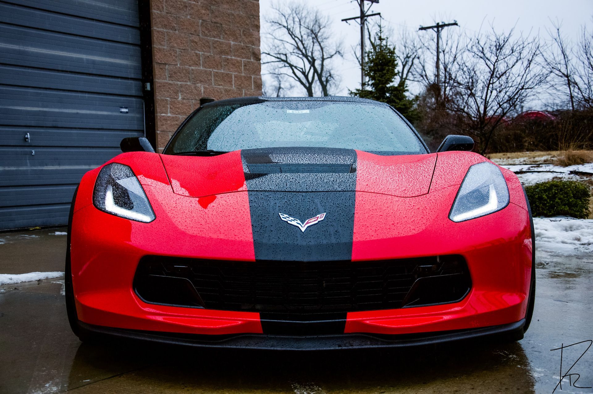 Red sports car with black racing stripe parked in front of a garage on a wet driveway
