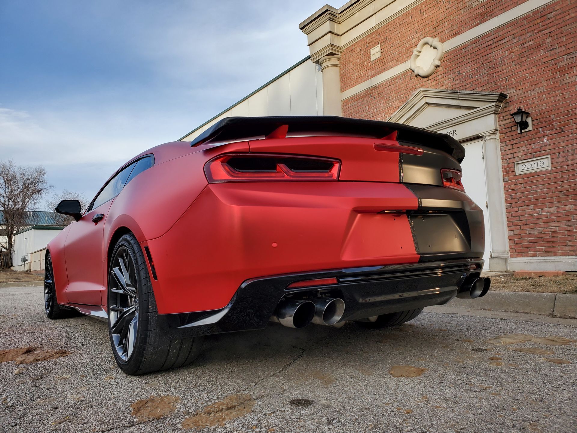 Red sports car parked beside a brick building, rear view on a gravel lot.