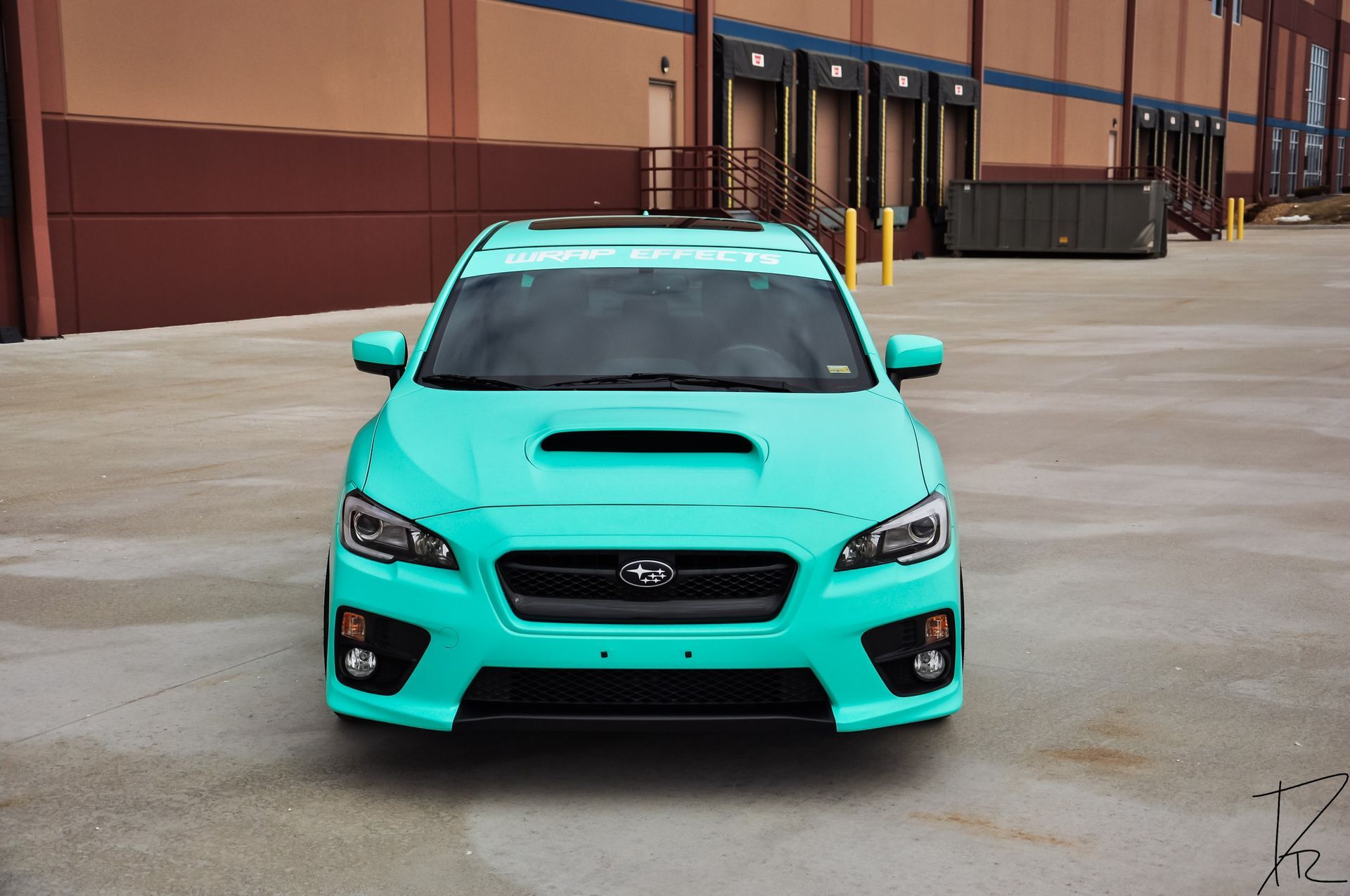 Turquoise sports sedan parked in an industrial lot, viewed from the front.