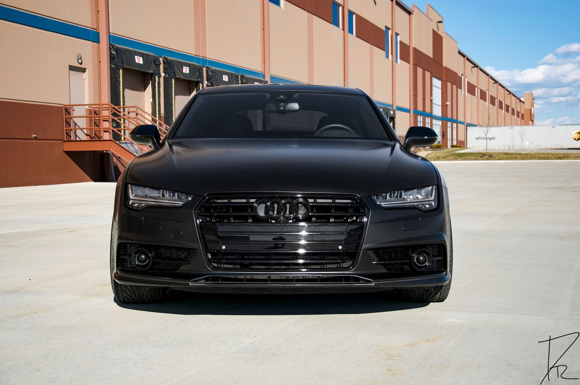 Black sedan parked in front of a tan industrial building on a sunny day