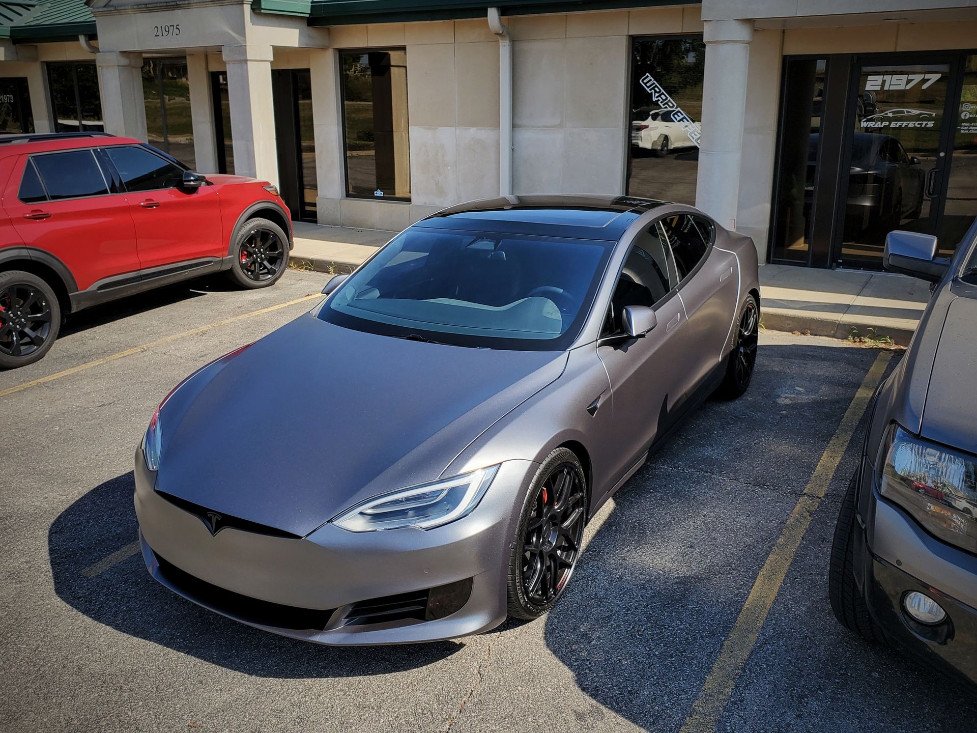 Gray Tesla sedan parked in a lot between a red SUV and a silver car outside storefronts