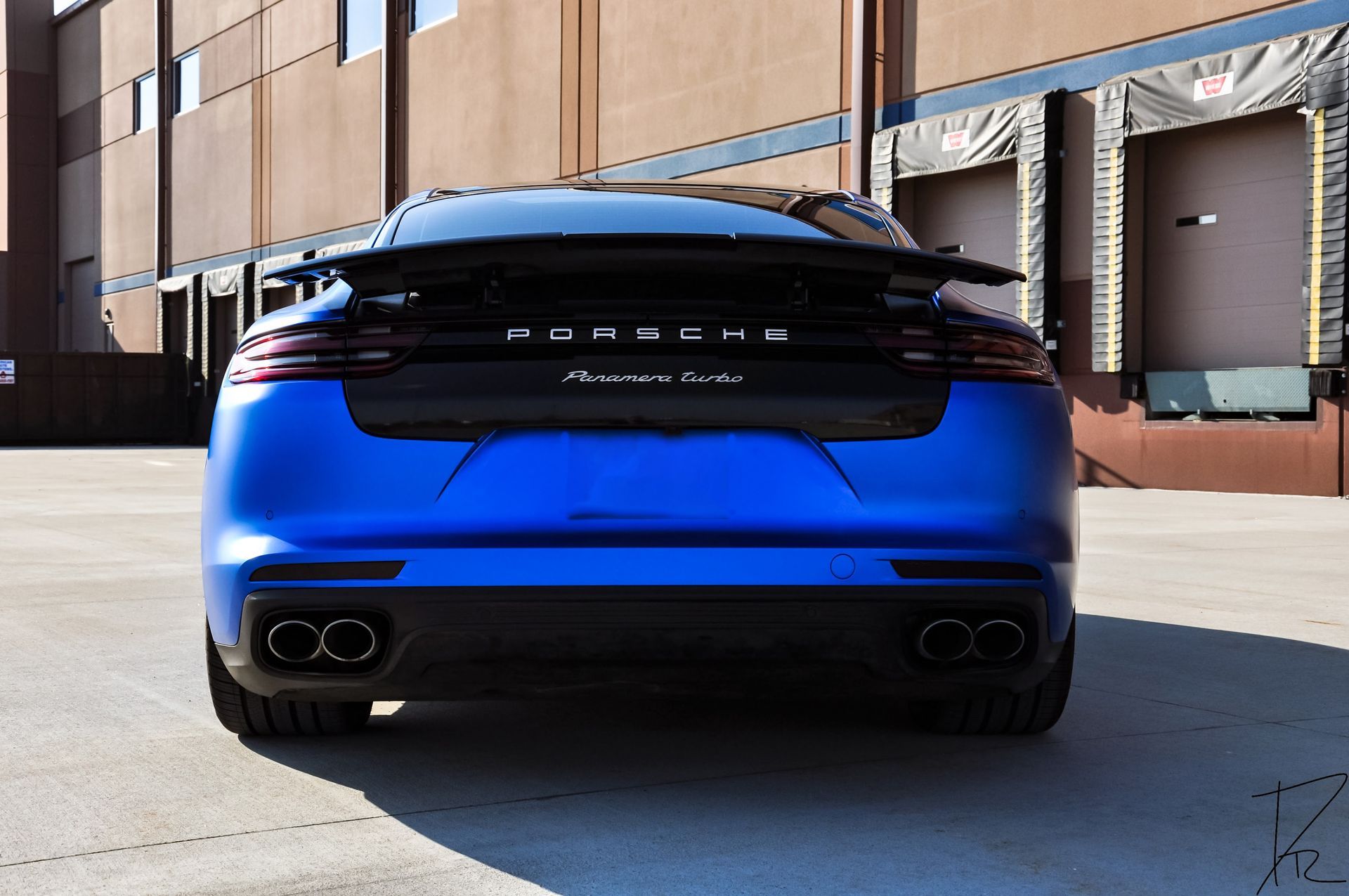 Blue Porsche rear view parked in a brick alley beside industrial doors