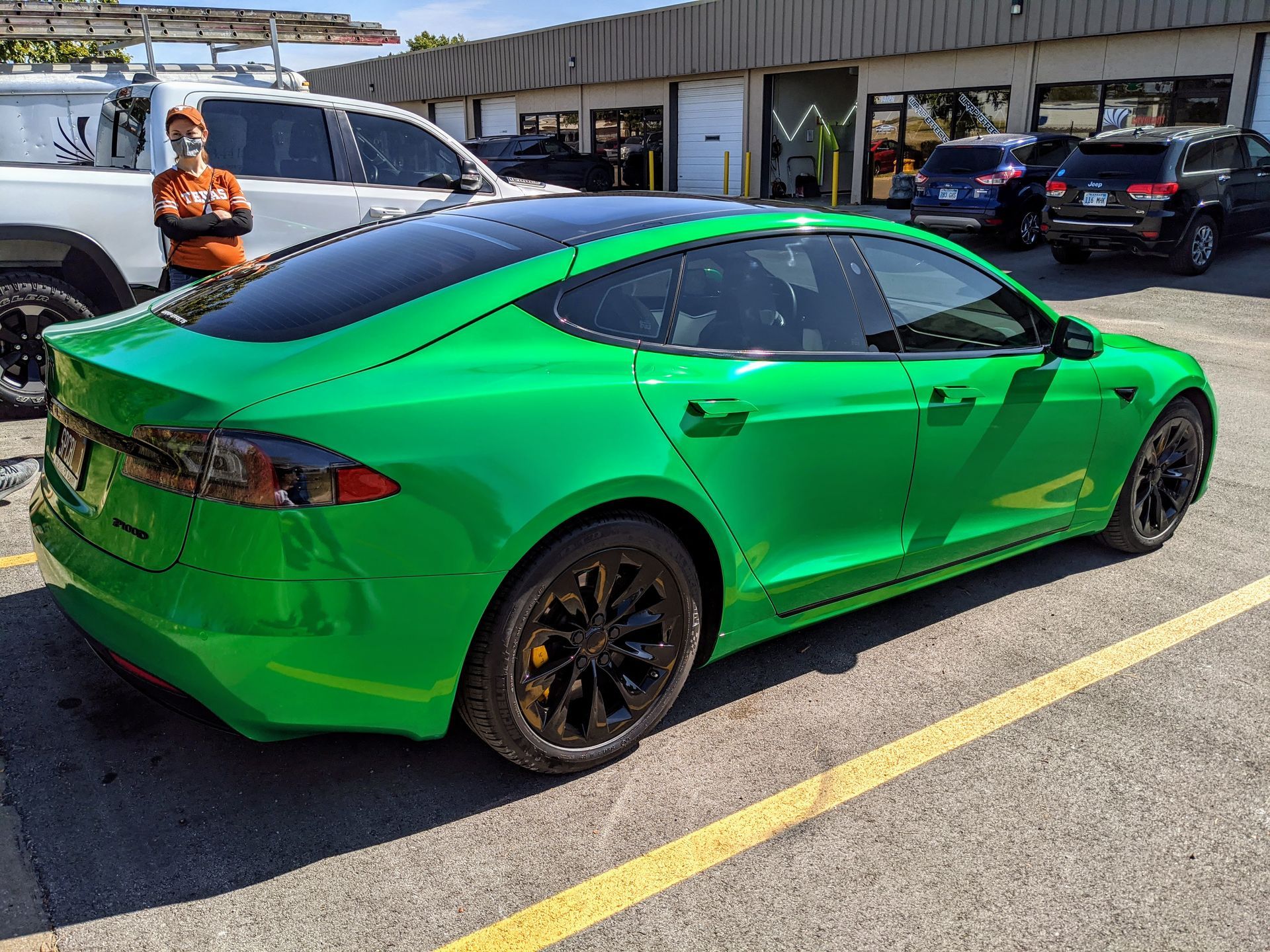 Green Tesla sedan parked in a lot, with a worker standing nearby and other cars in the background