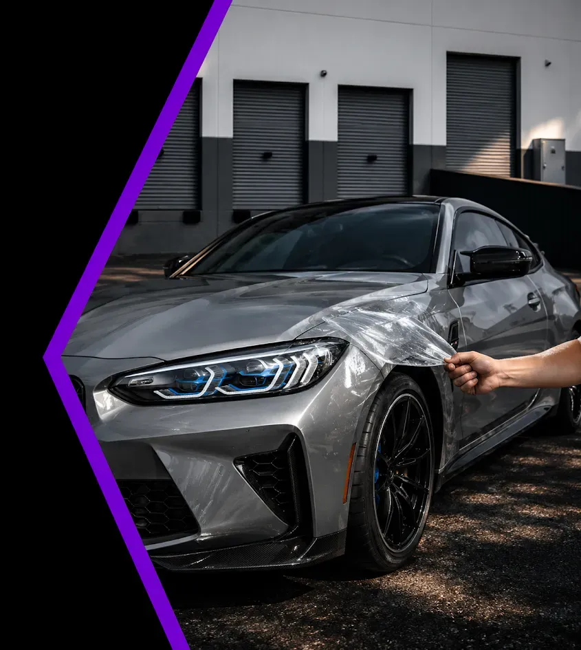 Silver sedan being wrapped in clear film outside a garage, with a purple diagonal border.