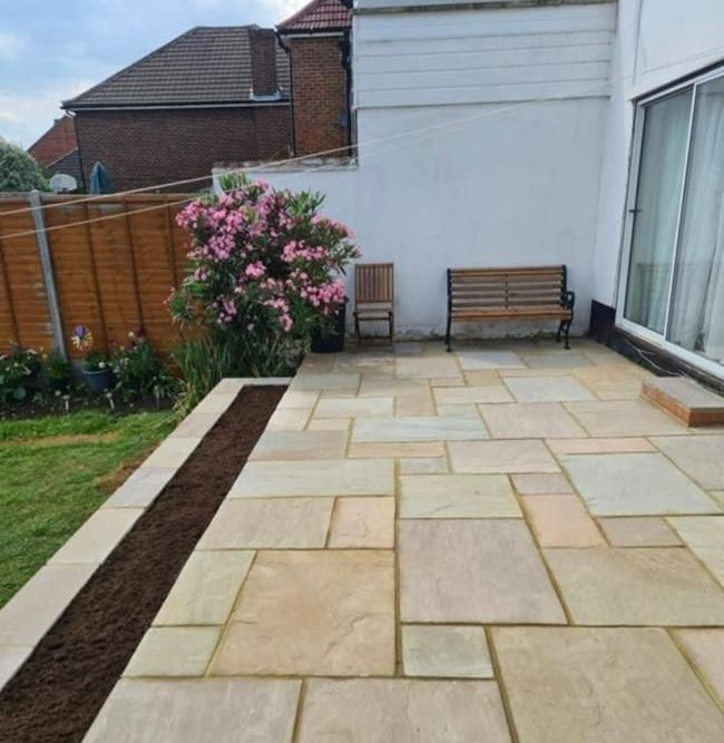 Paved patio with a bench, a pink flowering bush, and a long planter against a white wall.