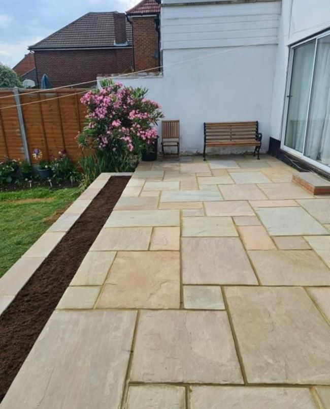 Patio with light-colored stone pavers, wooden bench and chair, next to a white wall and grassy lawn.